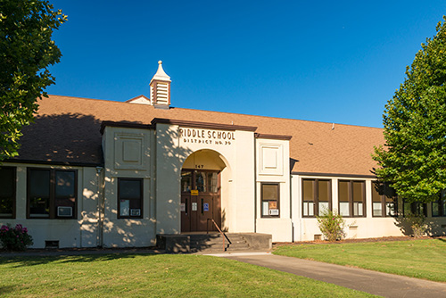 Exterior view of Riddle School, a single-story educational building with a central entrance flanked by windows. The name RIDDLE SCHOOL DISTRICT #70 is etched above the door.