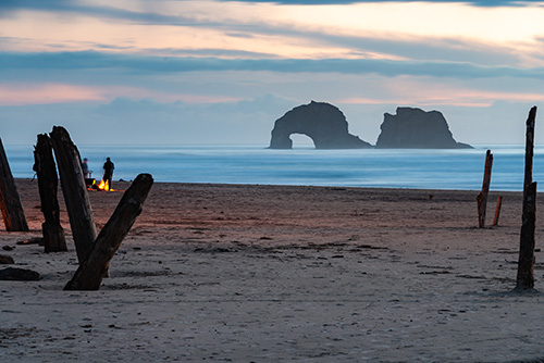 A beach scene at twilight with remnants of a wooden structure in the foreground and a distinctive rock formation visible in the ocean. A small figure stands near a glowing fire.