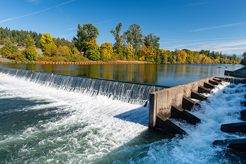 A river with a small waterfall created by a concrete barrier. The water is flowing vigorously over the barrier, creating white foam.