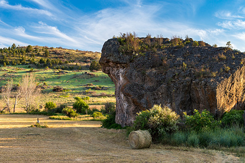 A scenic landscape featuring a large, rugged cliff with a flat top on the right side of the image. The cliff is covered with greenery at its summit and has steep, rocky sides.