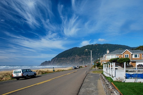 Cirrus clouds streak across blue sky above a 2 lane road running along side the beach. Ocean waves with white caps seen on left.