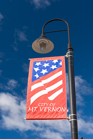 A red banner featuring a stylized American flag design with stars and stripes hangs from a black lamp post under a clear blue sky. The banner reads CITY OF MT. VERNON in white lettering.