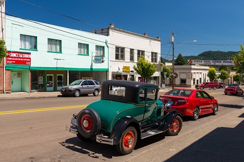 Street view of downtown Myrtle Creek. 2 lane road with businesses & sidewalks. A classic vintage car is parked by modern cars.