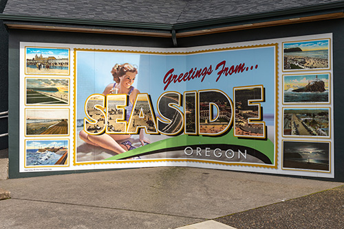 A mural reads Greetings from… SEASIDE OREGON in bold, stylized lettering. The mural features a collage of scenic views and landmarks within the letters, including a beach scene, rocky coastline, a lighthouse, and other picturesque landscapes typical of Seaside.