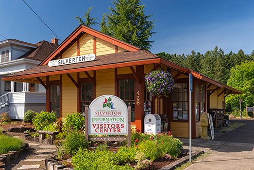 A small building with a bright orange roof and beige walls, labeled Silverton Information Visitors Center. The structure features a front porch with hanging purple flowers and is situated under a clear blue sky.