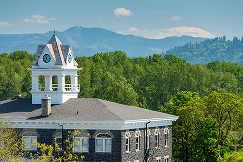 A building with a distinctive clock tower featuring a white cupola and weathervane. The building has a grey roof and is constructed with dark stone or brick. In the background, there are lush green trees and rolling hills.