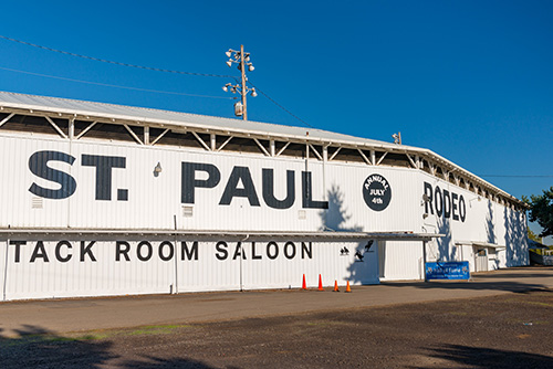 A large white building with ST. PAUL RODEO painted in black letters on its side, along with a logo featuring a bucking horse and rider silhouette. Below, there is a sign for TACK ROOM SALOON and two blue banners promoting the event.