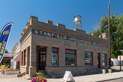 One-story stone building with sidewalk in front. Stanfield Irrigation District is printed on side. A watertower behind in distance.