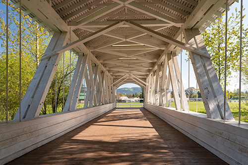 A wooden covered bridge with a truss design and a shingled roof. The interior shows the intricate wooden framework of the trusses, painted white, which supports the roof.