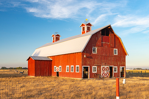 A traditional red barn with white trim under a clear blue sky. The barn features a large main structure with a pitched roof and two cupolas on top, and an attached smaller section with a similar roof style.