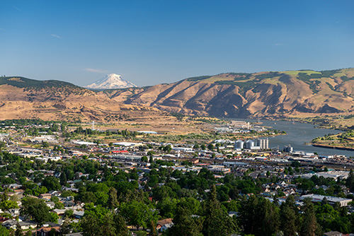 A panoramic view of a town nestled in a valley with a river running through it. The landscape features rolling hills and a prominent snow-capped mountain in the distance under a clear blue sky.