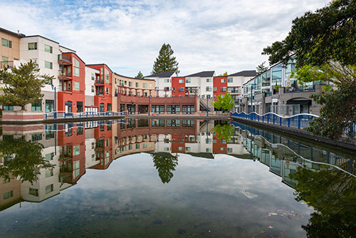 A calm and clear water body reflects a colorful urban landscape. The scene includes modern buildings with varied facades in shades of red, beige, and blue, featuring multiple windows and balconies. The buildings are arranged around the water, creating a symmetrical reflection.