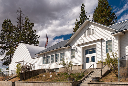 A white, single-story building with a gabled roof and a smaller adjoining section with an arched roof. The building has a blue door and multiple windows, some of which are open.