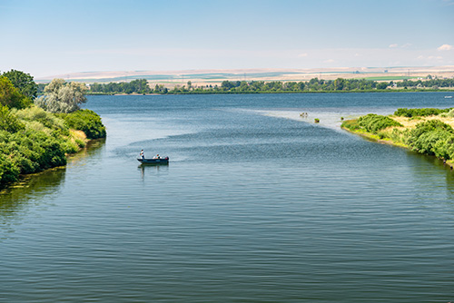 A serene river landscape with a small boat containing two individuals in the center, surrounded by lush greenery. The river is calm with gentle ripples on the water’s surface.