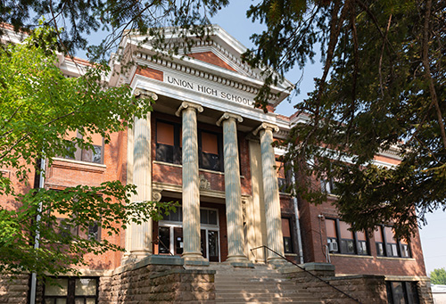 Exterior view of Union High School, featuring a classic red brick building with white trim. The school has a prominent set of stairs leading up to a grand entrance flanked by four large columns supporting an entablature with the school’s name inscribed.