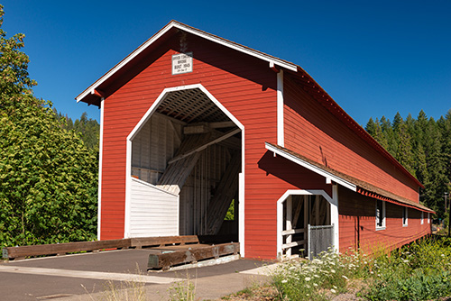 180 foot covered bridge using triple Howe Truss construction. A mix of evergreen & deciduous trees surround.