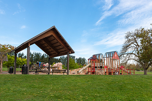 An outdoor park scene with a large wooden pavilion on the left, featuring a dark brown gabled roof and open sides. To the right is a colorful children’s playground with red, blue, and yellow equipment including slides and climbing structures.