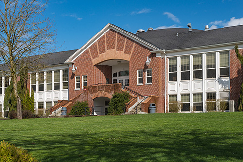 A traditional brick school building with a prominent central entrance featuring a set of stairs leading up to double doors under an arched doorway. The building has two wings with large, white-framed windows and is surrounded by a well-maintained lawn and several trees against a clear blue sky.