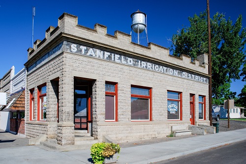 1-story stone building with sidewalk in front. "Stanfield Irrigation District" printed on side. A watertower behind in distance.