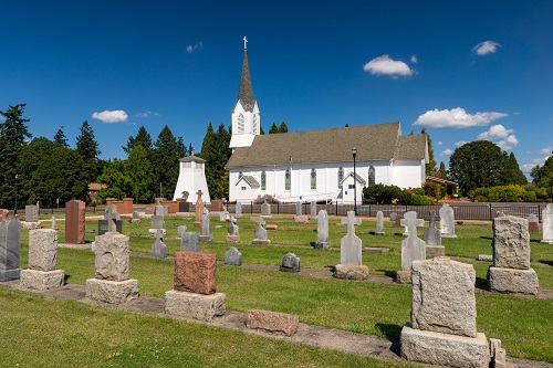 Single-story church with steeple in background. Foreground has neat rows of headstones for graves. Some have crosses for tops. 