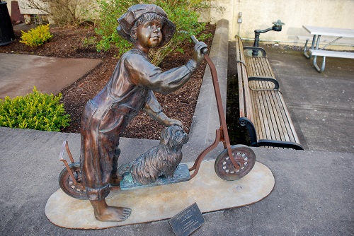 Bronze statue of boy with his dog & a scooter. The dog is small and sits on the foot board of the scooter. A bench is nearby.