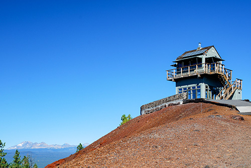 A multi-story wooden lookout tower perched atop a red-brown hill with a clear blue sky in the background.