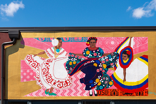 A vibrant mural painted on the side of a building, featuring three stylized figures in elaborate, patterned clothing.