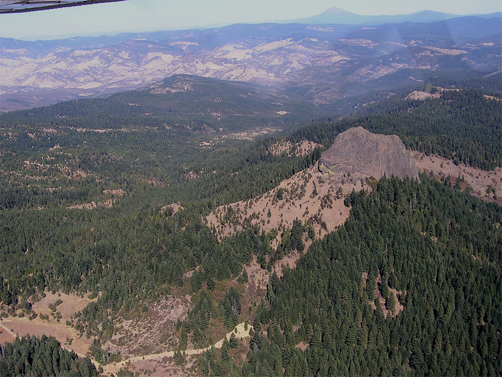 High‑elevation aerial view of a vast forested landscape with dense evergreen trees covering rolling hills. A large rocky outcrop rises from a partially tree‑covered ridge near the right side of the image.