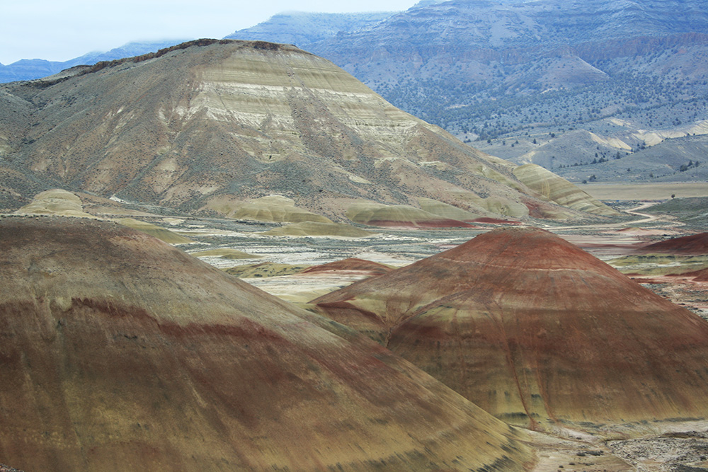 A landscape of rounded hills layered in shades of red, gold, and tan, forming the Painted Hills. The foreground shows smooth, gently sloping mounds with distinct horizontal color bands.