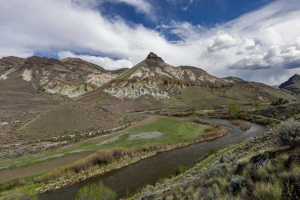 A wide landscape view of layered, multicolored hills and rocky formations rising behind a winding river. The hills display bands of tan, greenish-gray, and brown soil, with patches of low shrubs scattered across the slopes.