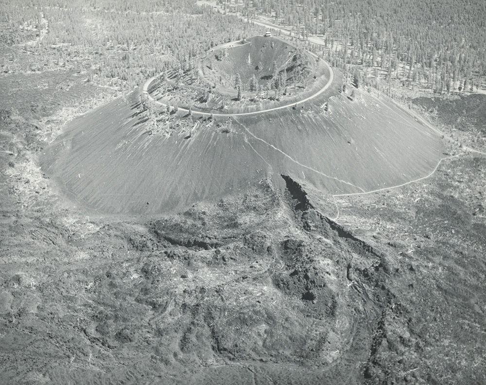 A black‑and‑white aerial photograph of a large volcanic cinder cone rising from a rugged landscape of hardened lava flows. The cone has steep, smooth slopes leading up to a circular crater rim at the top, with a narrow road or trail encircling the summit. 