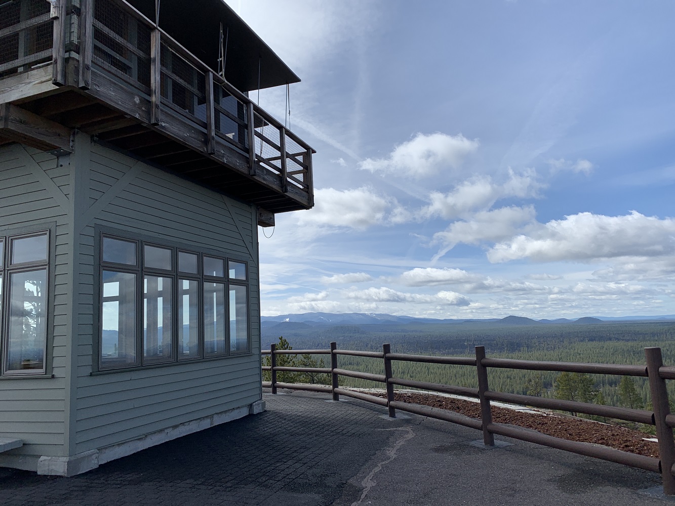 A fire lookout tower stands on the left side of the image, with light green siding and large windows beneath a wooden observation deck. A wooden railing lines the paved overlook in the foreground. 