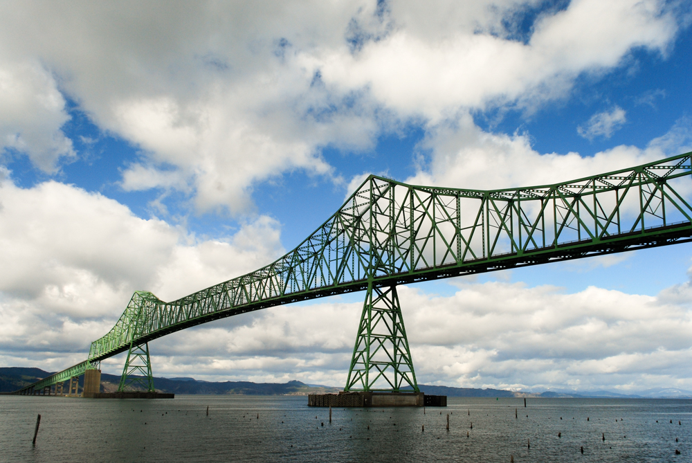 A wide green steel truss bridge stretches across a large body of water under a sky filled with scattered white and gray clouds.