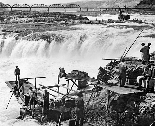 Native American men fish off platforms overhanging waterfalls.