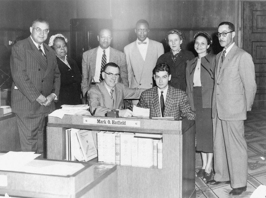 A black‑and‑white historical photograph showing a group of nine people standing and sitting around a desk in an office setting.