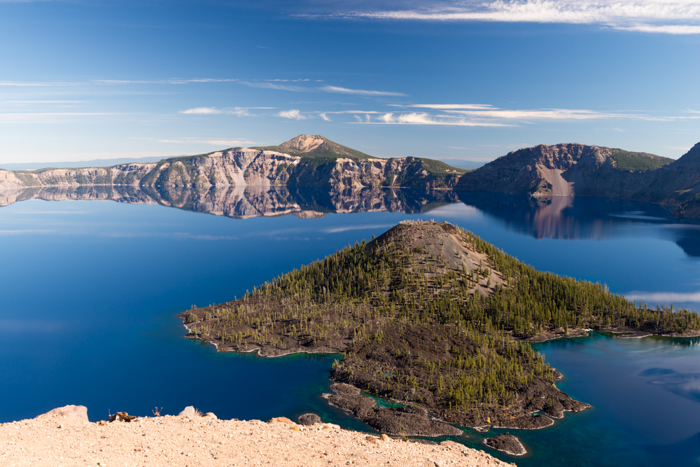 A wide scenic view of Crater Lake showing deep blue water surrounded by steep, rocky caldera walls. 