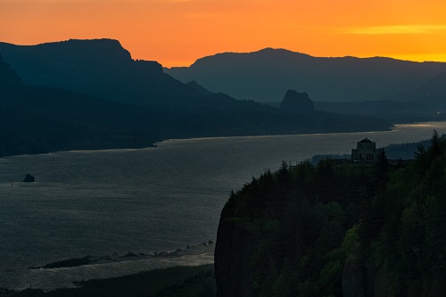 A scenic view of a wide river winding between forested cliffs and layered mountain ridges at sunset. The sky glows with deep orange light near the horizon, fading into darker tones above.