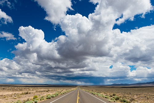 A long, straight highway stretches into the distance through a wide, open desert landscape. Low shrubs line the sides of the road, and a vast sky filled with large, billowing white clouds dominates the scene