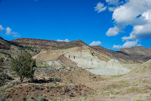 A landscape of layered, multi‑colored hills under a bright blue sky with scattered white clouds. 