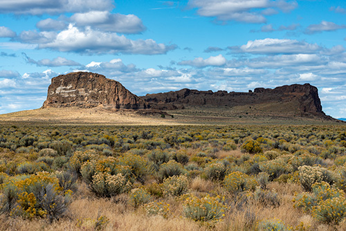 Fort Rock, a large circular tuff ring formation rising abruptly from a flat sagebrush plain in central Oregon. Its steeply sloping walls display steep cliffs composed of light-gray volcanic tuff, with a roughly 400-foot diameter inner crater. 