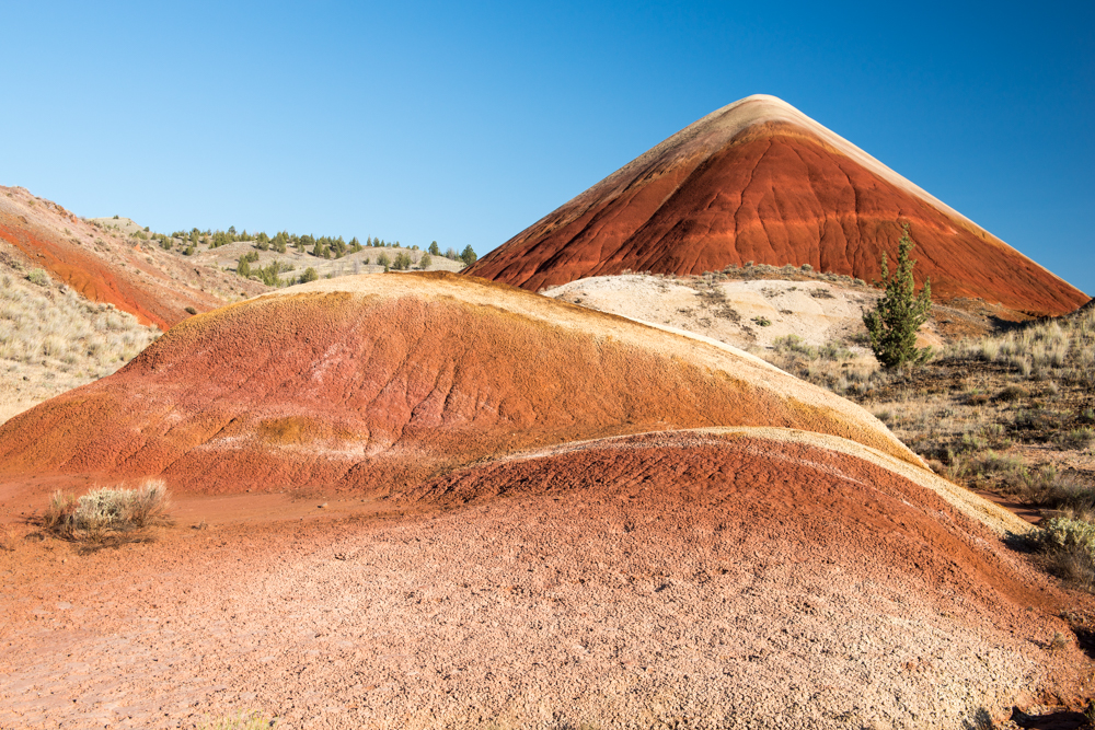 A landscape of gently sloping, multicolored hills featuring vivid bands of red, orange, yellow, and tan earth. In the background, a tall, cone‑shaped hill rises with deep red and brown layers.