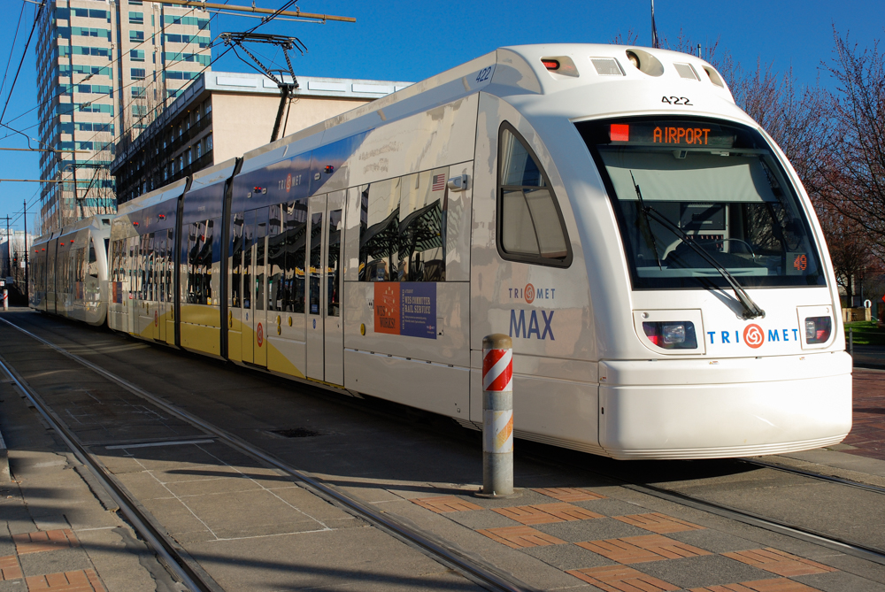 A modern white light‑rail train labeled TriMet MAX is stopped at a station platform. The destination sign on the front reads Airport.
