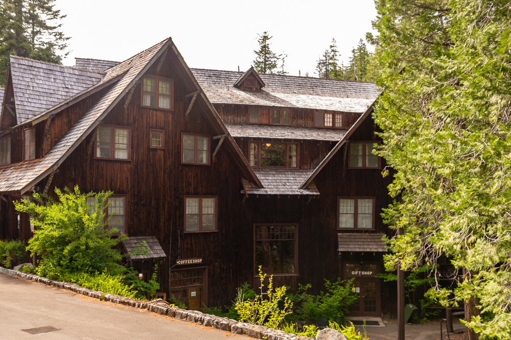 A large, rustic wooden lodge with dark brown siding and multiple steep gabled roofs sits among tall evergreen trees.