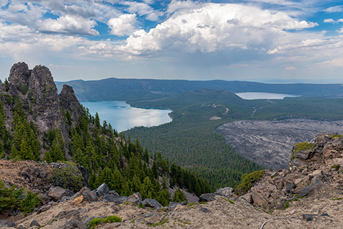 A high‑elevation view overlooking a large forested landscape with two blue lakes in the distance. Jagged volcanic rock formations rise in the foreground beside a slope covered in evergreen trees.