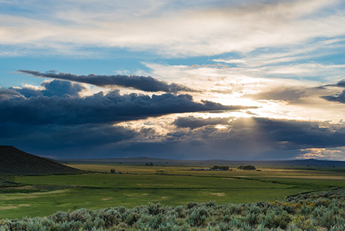 A scenic landscape. A vast grassland with a gentle slope on the left. Above, a dramatic sky unfolds, where sunlight pierces through an opening in the heavy cloud cover.