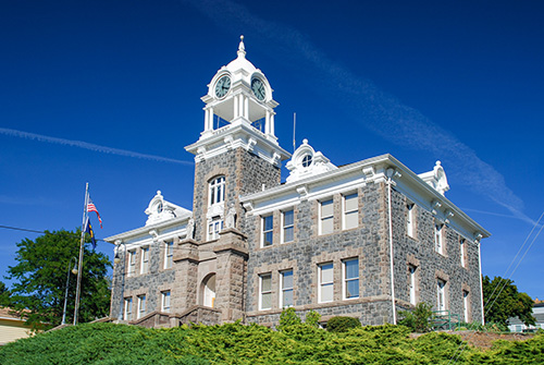 Front façade of the Morrow County Courthouse in Heppner, Oregon — a two-story dark basalt stone building with lighter trim, featuring a central clock tower topped by a domed cupola