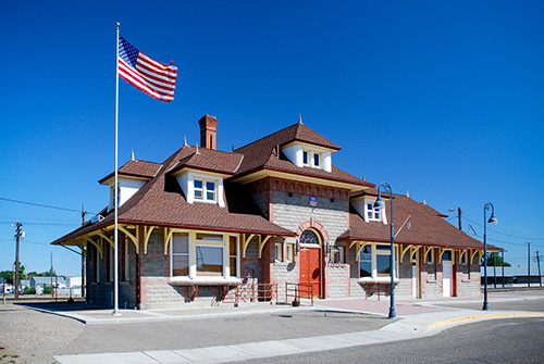 A historic brick train station with an American flag waving in the clear blue sky. The station features a red-tiled roof, yellow