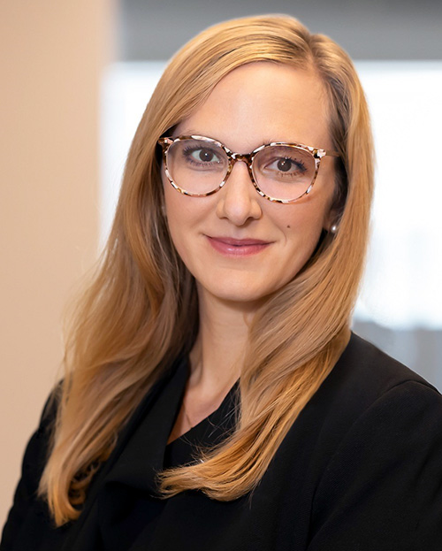 Christina Stephenson has long blonde hair and is wearing glassess and a black suit.
