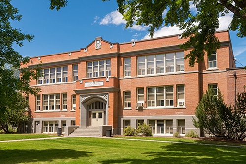A two-story brick building with large windows and a central entrance featuring a set of concrete stairs leading up to double doors.