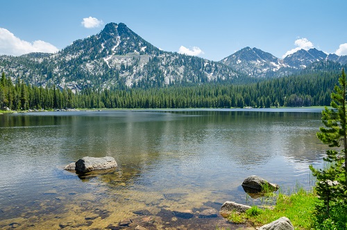 Lake boardered by fir trees with 3 mountain peaks in back. Mountains have slight dusting of snow.